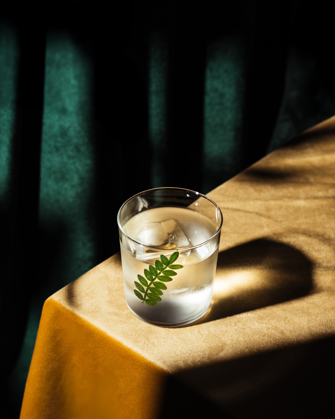 A clear drink with ice and a green leaf garnish is elegantly served in a glass on a soft golden tablecloth